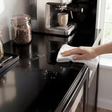 Person cleaning a black kitchen counter with a white cloth, next to a coffee machine and jar of coffee beans.