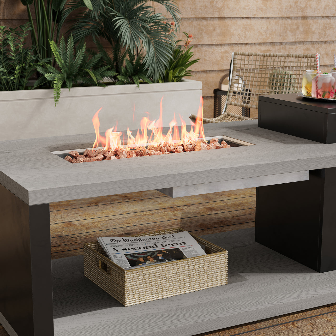 Outdoor fire pit table with flames, newspaper, and plants in the background.