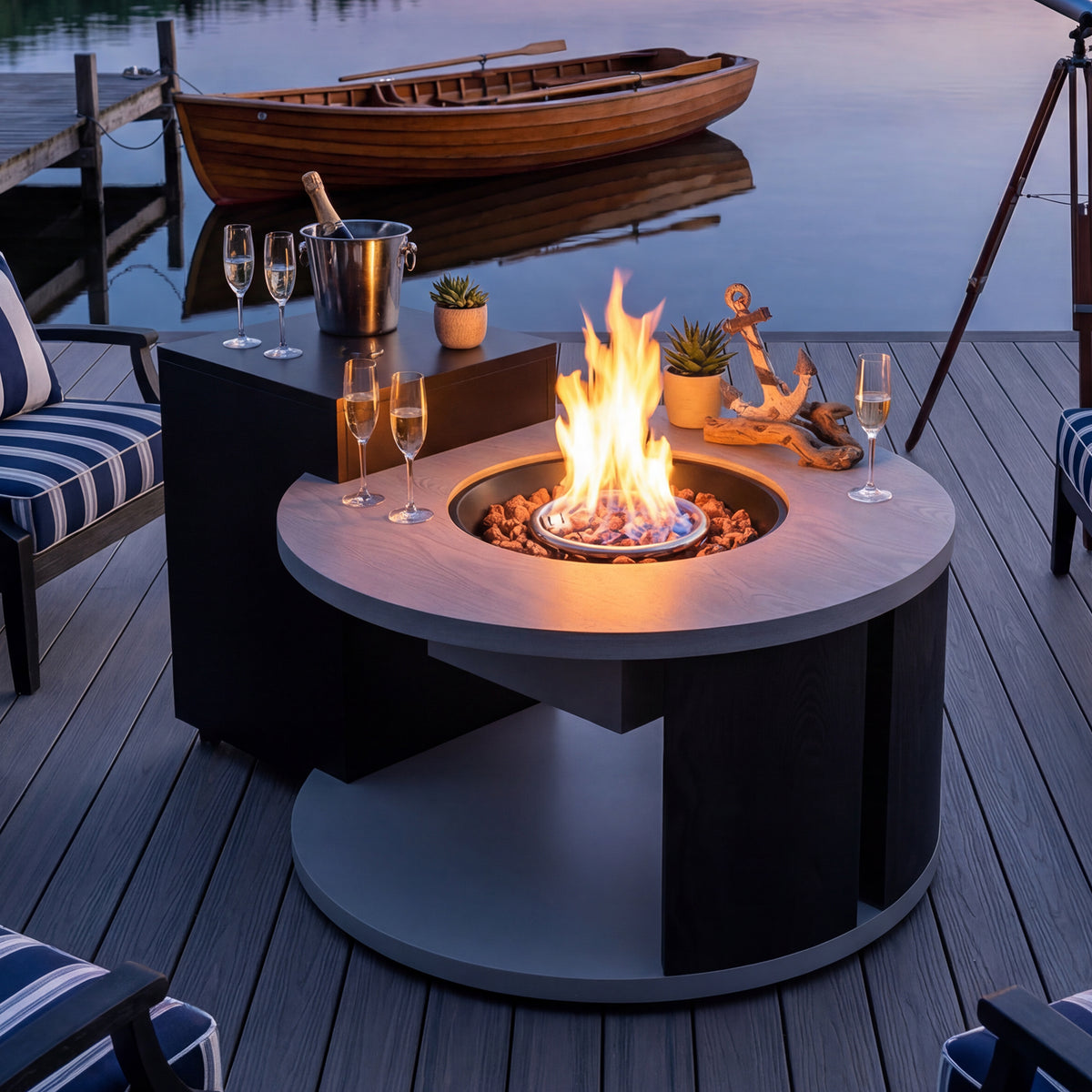 Fire pit table with flames on a wooden deck by a body of water, with chairs and decorative items around.