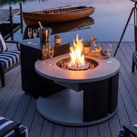Fire pit table with flames on a wooden deck by a body of water, with chairs and decorative items around.