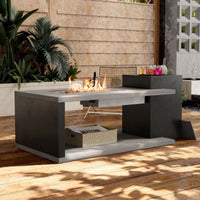 Modern outdoor fire pit table with flames on a wooden deck, surrounded by plants and a building.