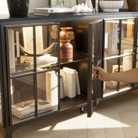 Hand opening a glass door of a decorative cabinet with books and decor items inside.
