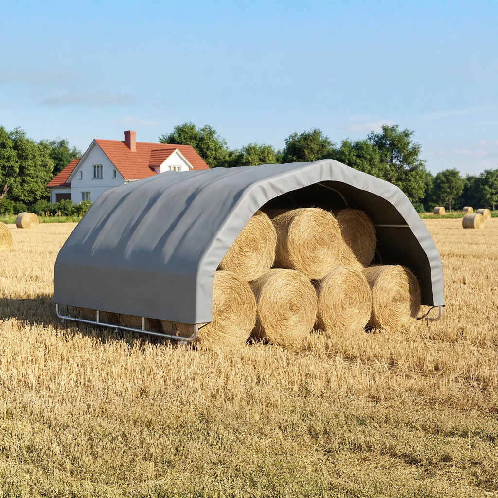 Hay bales stacked inside a gray portable shelter in an open field with a house in the background