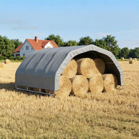 Hay bales stacked inside a gray portable shelter in an open field with a house in the background